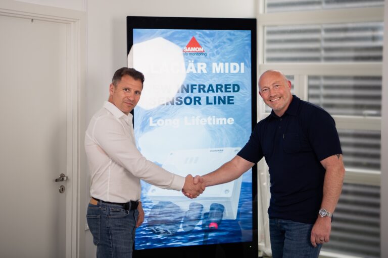 Two men in casual shirts shake hands beside a tall digital display with a blue water background and white text, in a bright office setting.