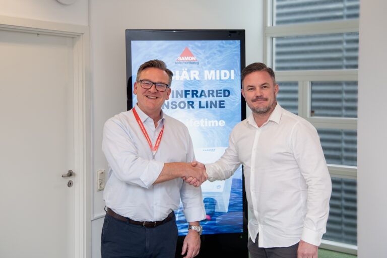Two professionals in white shirts shake hands in an office, with a tall blue branding display behind them reading SAMON safe monitoring.”