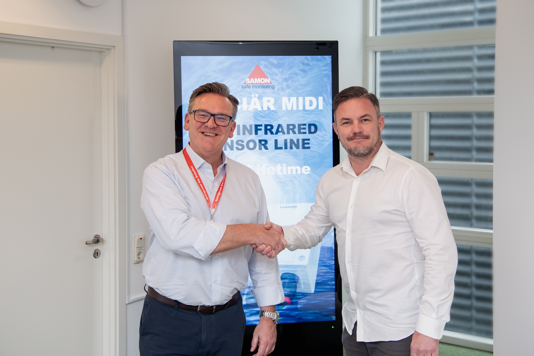 Two professionals in white shirts shake hands in an office, with a tall blue branding display behind them reading SAMON safe monitoring.”