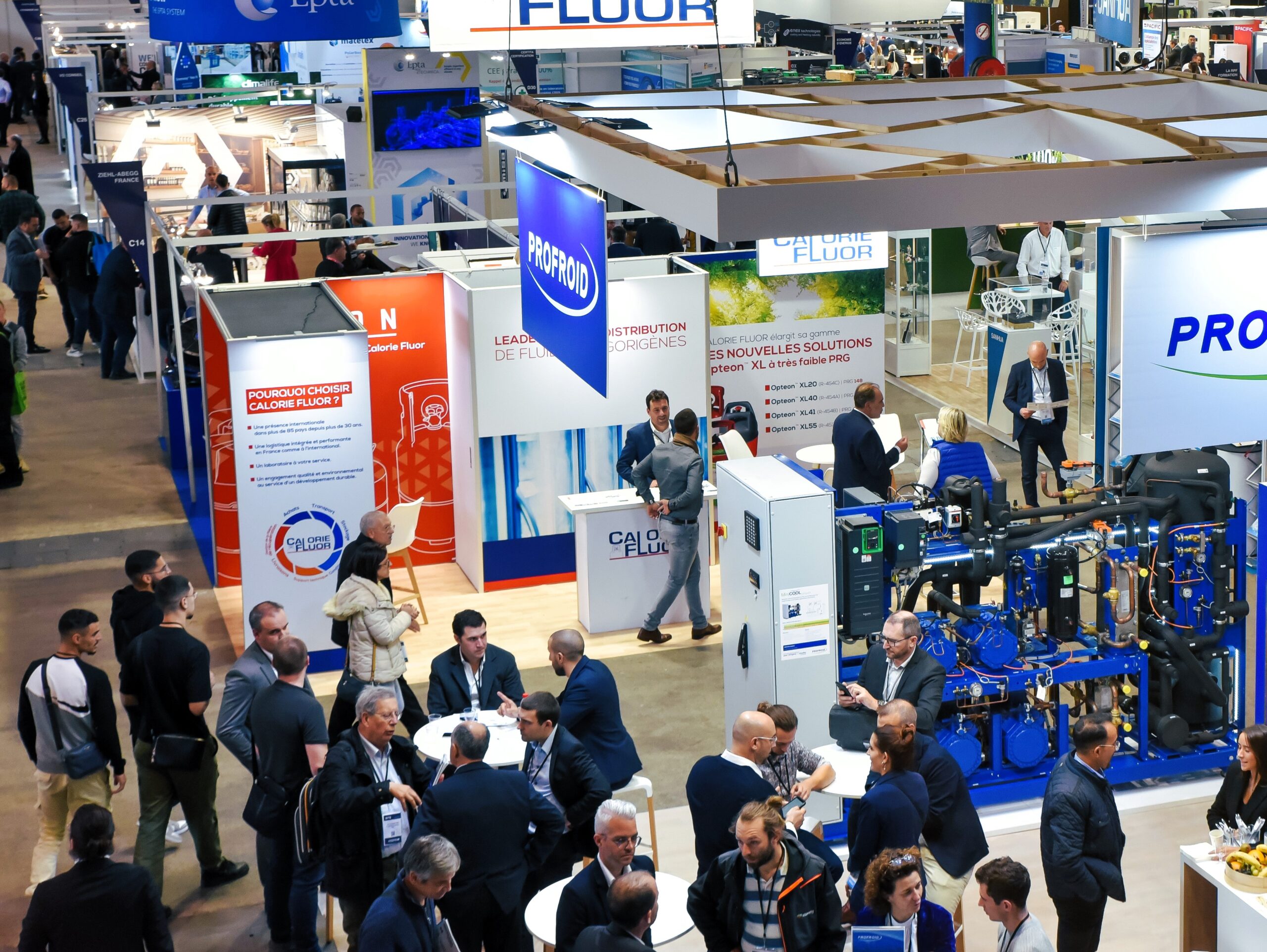 Busy trade show hall with numerous exhibition booths, banners, and groups of professionals networking around white tables.