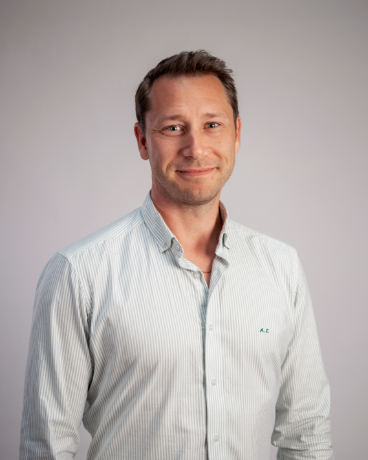Portrait of a smiling man in a light striped button-down shirt against a neutral gray background.