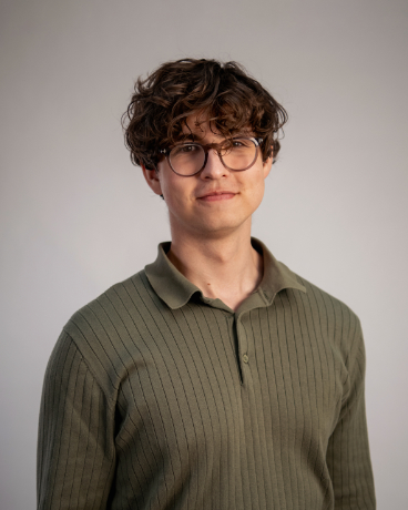 Young man with curly brown hair and glasses wearing a green ribbed polo shirt, standing against a light gray backdrop.