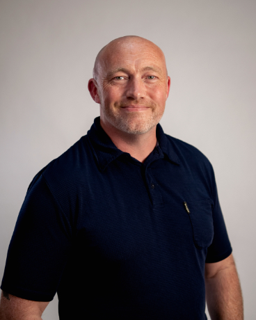 Smiling bald man wearing a navy polo shirt posing against a light gray studio background.