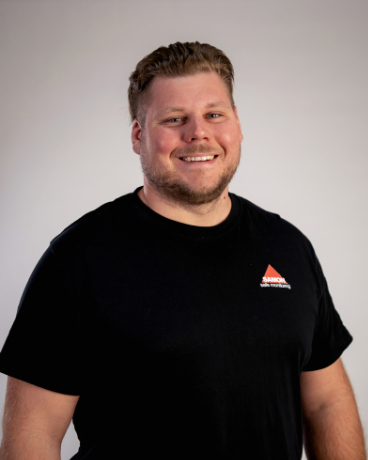 Smiling man in a black T-shirt with a small logo on the chest, standing against a neutral gray background.