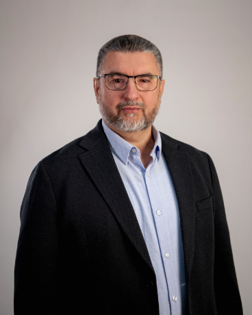 Professional portrait of a middle-aged man with gray hair and beard wearing glasses, a blue shirt, and a black blazer.
