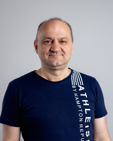 Retrato de um homem de meia-idade com cabelo curto rapado vestindo uma t-shirt azul-marinho com texto branco vertical ao longo da zona direita do peito, de frente para a câmara.