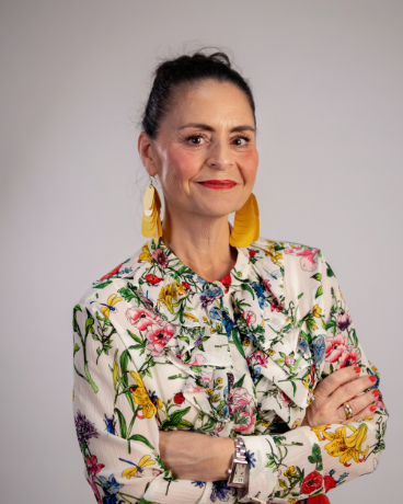 Portrait of a smiling woman with dark hair in a bun, wearing a colorful floral blouse and bold yellow earrings, arms crossed.