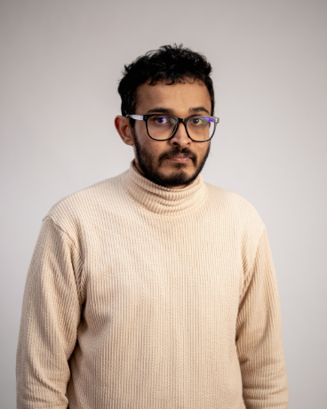 Portrait of a man with dark hair, beard and glasses wearing a cream knit turtleneck against a light gray background.