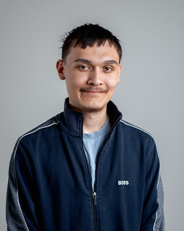 Retrato de um jovem com cabelo escuro curto vestindo um casaco azul-marinho com fecho de correr sobre um fundo cinzento, sorrindo subtilmente.
