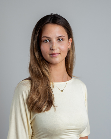 Jovem mulher com cabelo castanho comprido vestindo um top amarelo pálido, olhando para a câmara sobre um fundo cinzento.