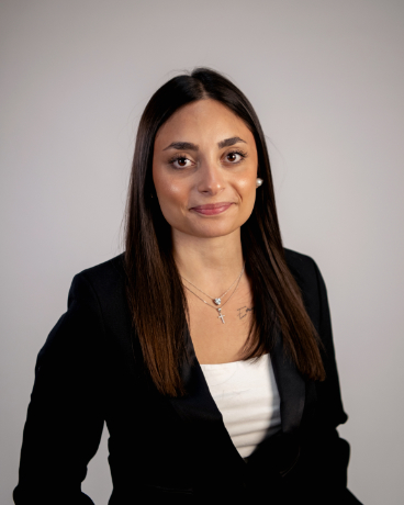 Professional headshot of a woman in a black blazer and white top, with straight dark hair and a subtle smile, neutral background.