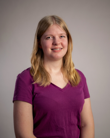 Young woman with shoulder-length blonde hair, wearing a purple V-neck shirt, smiling at the camera against a gray studio backdrop.