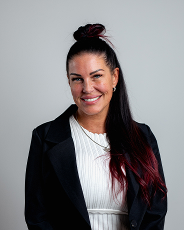 Smiling woman with dark hair in a bun, wearing a white knit sweater and black blazer, against a light gray background.
