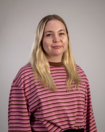 Portrait of a woman with long blonde hair, wearing a pink striped sweater, smiling at the camera.