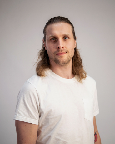 Portrait of a man with shoulder-length hair wearing a white t-shirt, standing against a light gray background and looking at the camera.