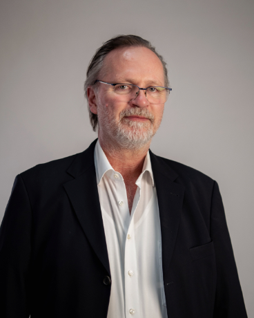Portrait of a middle-aged man with gray hair and beard, wearing glasses, a white shirt, and a black blazer, looking at the camera.