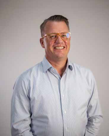 Professional headshot of a smiling man in a light blue button-down shirt and amber glasses against a neutral gray background.